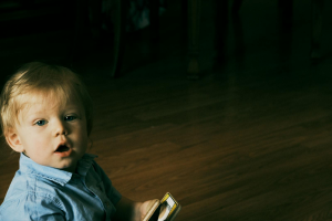 A young child curiously holding a smartphone while sitting on a wooden floor indoors.